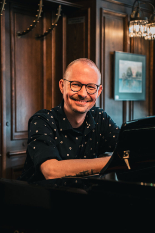 Ein Mann mit Brille sitzt am Klavier und lächelt