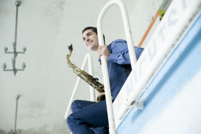 Ein Mann mit einem Saxophon sitzt am Beckenrand und lächelt