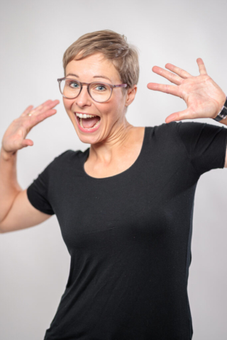 Brustbild einer Frau mit Brille in schwarzem Shirt die die Hände neben Dem Kopf mit gespreizten Fingern hoch hält und lacht