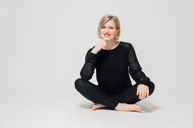 Eine Frau sitzt im Schneidersitz barfuß auf weißem Boden, die rechte Hand hat sie am Kinn und lächelt