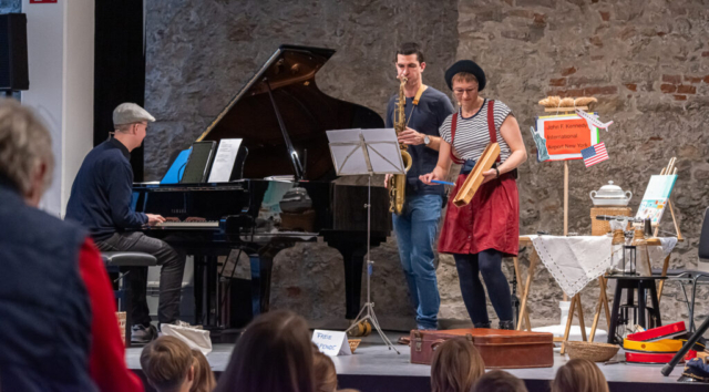 Katharina Osztovics, Markus Osztovics und Philipp Eder musizieren auf der Bühne mit Guiro, Saxophon und Klavier